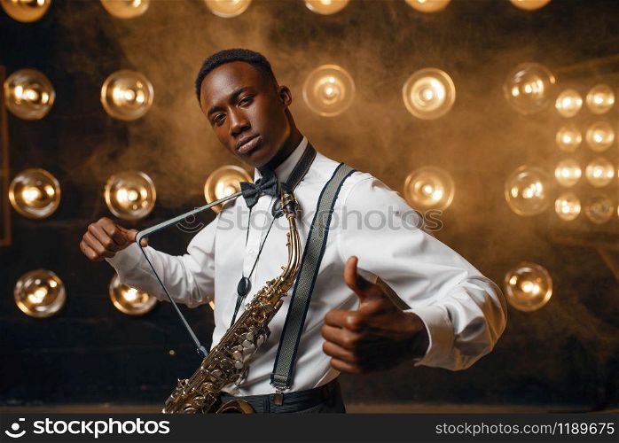 Black smiling jazz performer with saxophone on the stage with spotlights. Black jazzman preforming on the scene. Smiling jazz performer with saxophone on stage
