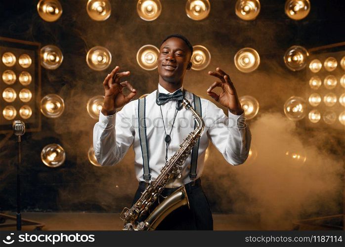 Black smiling jazz performer with saxophone shows OK sign on the stage with spotlights. Black jazzman preforming on the scene. Black jazz performer with saxophone shows OK sign