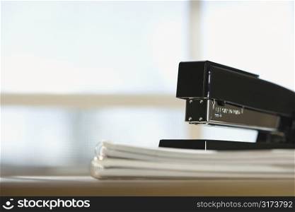 Black stapler on stack of paper with window in background.
