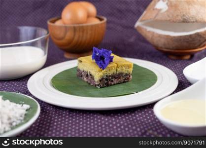 Black sticky rice and custard on a banana leaf in a white plate with butterfly pea flowers.
