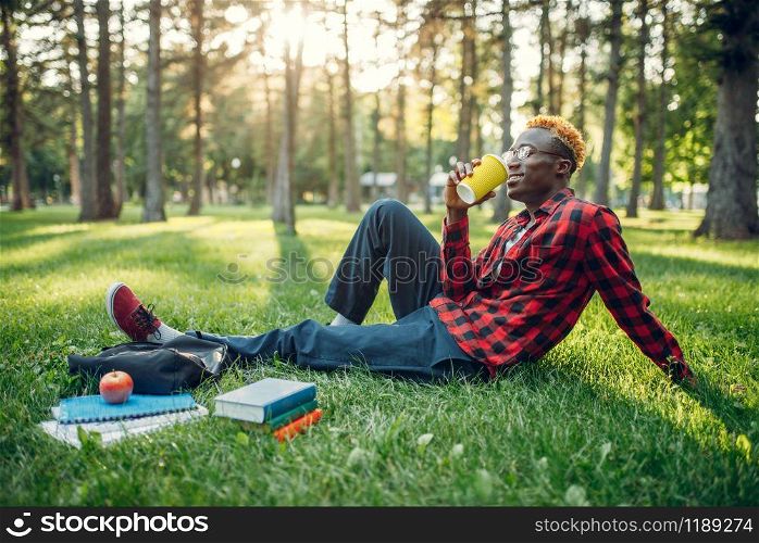 Black student drinks coffee on the grass in summer park. A teenager studying outdoors and having lunch. Black student drinks coffee on the grass in park