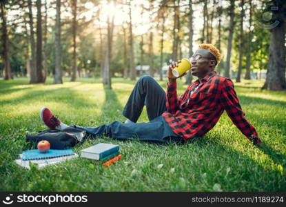 Black student drinks coffee on the grass in summer park. A teenager studying outdoors and having lunch. Black student drinks coffee on the grass in park