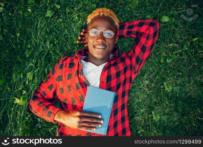 Black student in glasses reading book on the grass in summer park. A teenager studying outdoors and having lunch. Black student in glasses reading book on the grass