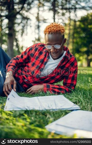 Black student in glasses reading book on the grass in summer park. A teenager studying outdoors and having lunch. Black student in glasses reading book on the grass