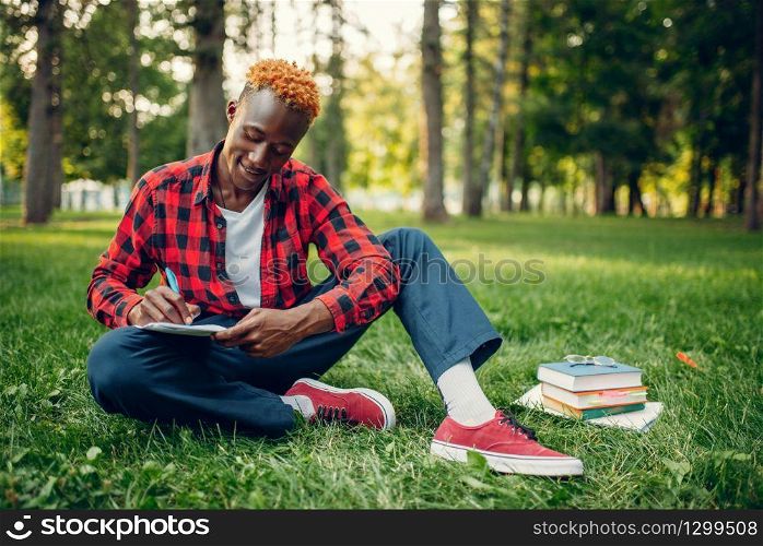 Black student in glasses writing in notebook on the grass in summer park. A teenager studying outdoors and having lunch. Black student writing in notebook on the grass