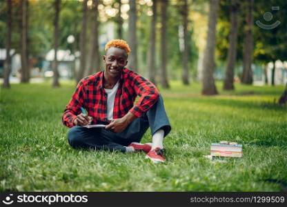 Black student in glasses writing in notebook on the grass in summer park. A teenager studying outdoors and having lunch. Black student writing in notebook on the grass