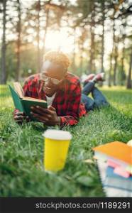 Black student in headphones lying on the grass and reading a book in summer park. A teenager studyingand leisures outdoors. Black student in headphones lying on the grass