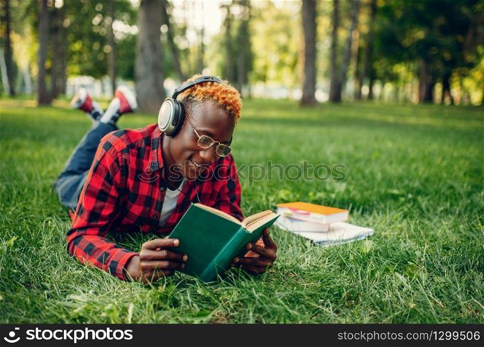 Black student in headphones lying on the grass and reading a book in summer park. A teenager studyingand leisures outdoors. Black student in headphones lying on the grass