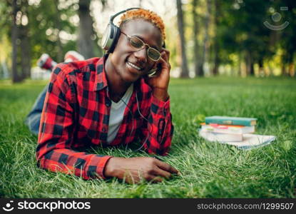 Black student in headphones resting on the grass in summer park. A teenager studying and leisures outdoors. Black student in headphones resting on the grass