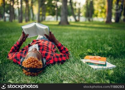 Black student in headphones resting on the grass in summer park, top view. A teenager studying and leisures outdoors. Black student resting on the grass, top view