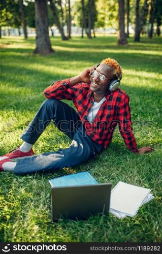 Black student in headphones sitting on the grass in summer park. A teenager studying and leisures outdoors. Black student in headphones sitting on the grass