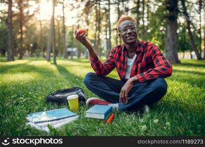 Black student with apple sitting on the grass in summer park. A teenager studying outdoors and having lunch. Black student with apple sitting on the grass