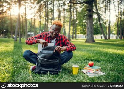 Black student with backpack sitting on the grass in summer park. A teenager studying outdoors and having lunch. Black student with backpack sitting on the grass