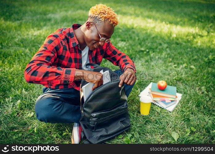 Black student with backpack sitting on the grass in summer park. A teenager studying outdoors and having lunch. Black student with backpack sitting on the grass