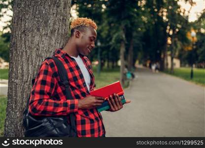 Black student with book and backpack standing near the tree in summer park. A teenager studying outdoors and having lunch. Black student with book and backpack in park