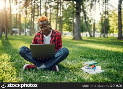 Black student with laptop sitting on the grass in summer park. A teenager studying and leisures outdoors. Black student with laptop sitting on the grass