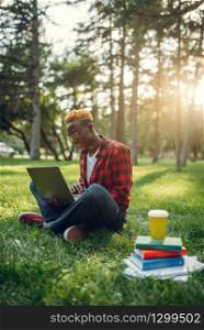 Black student with laptop sitting on the grass in summer park. A teenager studying and leisures outdoors. Black student with laptop sitting on the grass