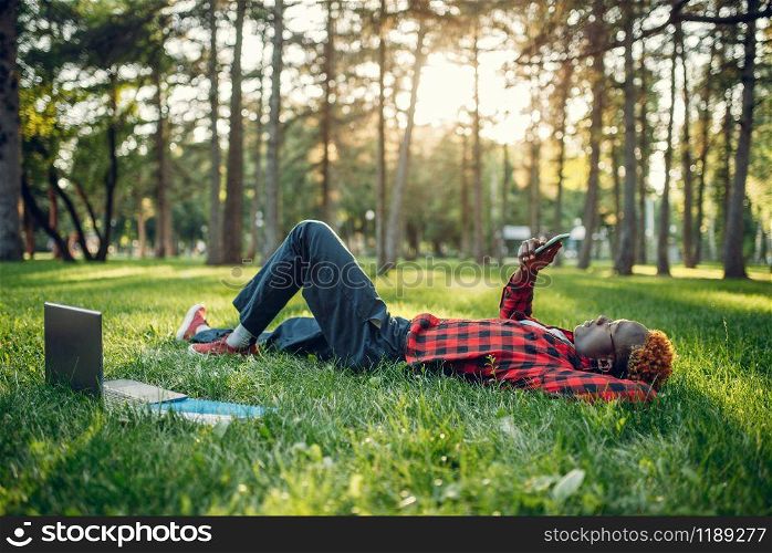 Black student with phone lying on the grass in summer park. A teenager studying and leisures outdoors. Black student with phone lying on the grass