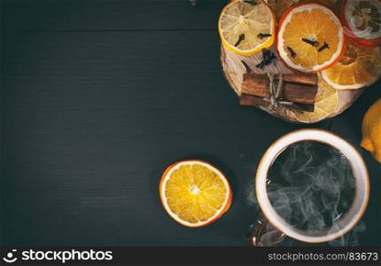 Black tea in a mug next to slices of dried orange and cinnamon on a black wooden surface, top view