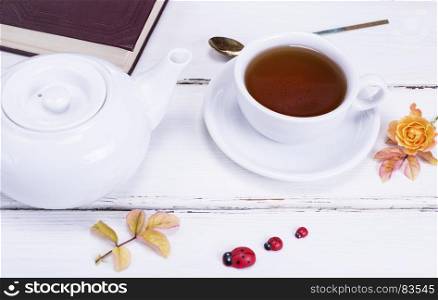 black tea in a round white cup with a saucer, next to a white tea pot and a closed book