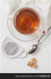 Black tea tea bag on ceramic plate with glass of tea and strainer infuser and cane sugar on white background.