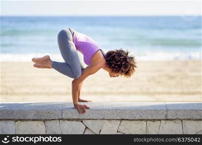 Black woman, afro hairstyle, doing yoga in the beach. Young Female wearing sport clothes with sea at the background