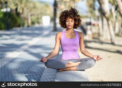 Black woman, afro hairstyle, doing yoga in the beach with eyes closed. Young Female wearing sport clothes in lotus figure with defocused background.