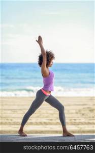 Black woman, afro hairstyle, doing yoga in warrior asana in the beach. Young Female wearing sport clothes with sea at the background