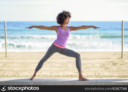 Black woman, afro hairstyle, doing yoga in warrior pose in the beach. Young Female wearing sport clothes with sea at the background