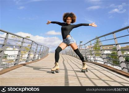 Black woman, afro hairstyle, on roller skates riding outdoors on urban bridge with open arms. Smiling young female rollerblading on sunny day. Beautiful clouds in the sky.