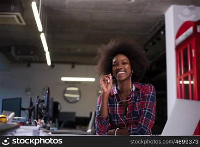 black woman in modern office at her workplace speeking on phone over earphones and working on laptop computer