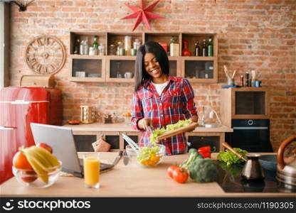 Black woman looking recipe on laptop, young ethnic girl cooking on the kitchen. African female person preparing vegetable salad. Black woman looking recipe on laptop