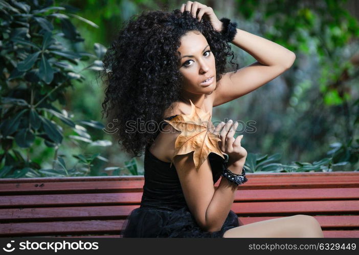 Black woman with a dry leaf in an urban park. Beautiful female with afro hairstyle. Young girl wearing autumn clothes.