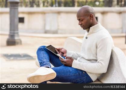 Black young man looking at tablet computer and smartphone in urban background. Young african guy with shaved head wearing casual clothes.