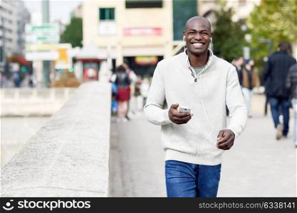 Black young man with a smartphone in his hand in urban background. Young african guy with shaved head wearing casual clothes and white headphones.