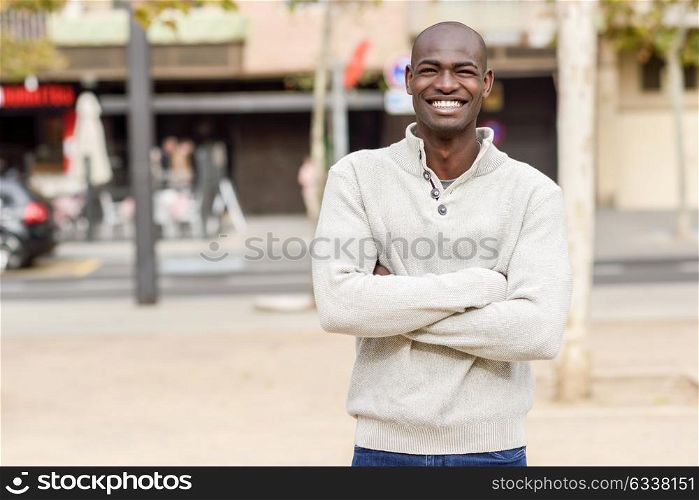 Black young man with arms crossed smiling in urban background. Young african guy with shaved head wearing casual clothes.
