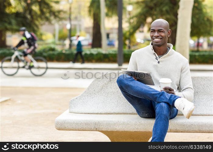 Black young man with tablet computer and take away coffee in urban background. Young african guy with shaved head wearing casual clothes.