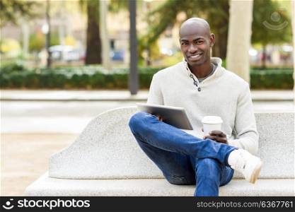 Black young man with tablet computer and take away coffee in urban background. Young african guy with shaved head wearing casual clothes.