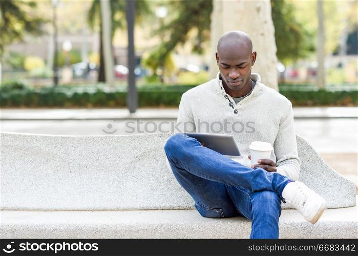 Black young man with tablet computer and take away coffee in urban background. Young african guy with shaved head wearing casual clothes.. Black young man with tablet computer and take away coffee