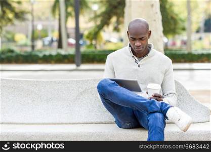 Black young man with tablet computer and take away coffee in urban background. Young african guy with shaved head wearing casual clothes.. Black young man with tablet computer and take away coffee