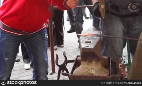 blacksmith holds metallic rod on anvil and young guy strikes big hammer flattened it