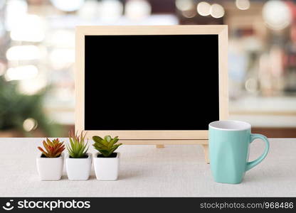 Blank chalkboard and green coffee cup standing on sack tablecloth over blur restaurant with bokeh background, space for text, mock up, product display montage