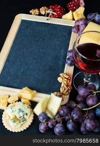 Blank chalkboard surrounded by glass of red wine, various types of cheese, walnuts and grapes on a black background