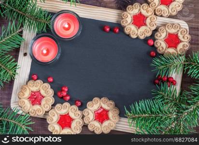 Blank chalkboard surrounded by green fir branches, two red burning candles and few cookies with berry jam, top view, flat lay