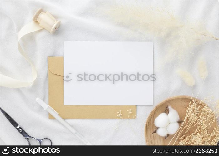 Blank greeting card invitation Mockup 5x7 on envelope with dry flowers and ribbon on white fabric background, flat lay, mockup