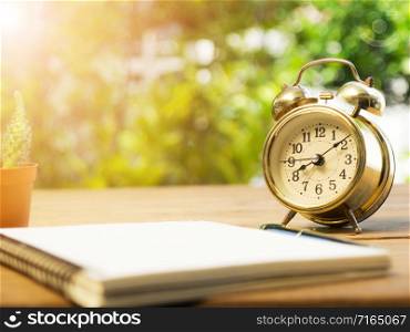 Blank notebook with vintage alarm clock and black pen and cactus on wood table. The background is green from tree and light bokeh