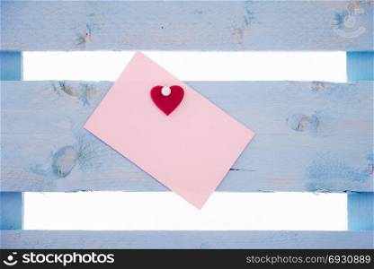 Blank pink paper note with a red heart glued to it and together pinned to a blue wooden fence.