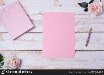 Blank pink paper sheet mockup with notebook, pen and roses on the white wooden table