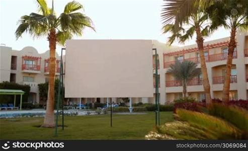 Blank presentation screen on summer resort standing between palms trees