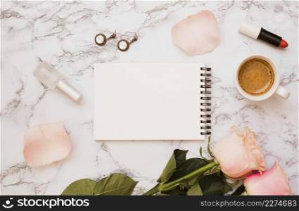 blank spiral notepad with earrings nail varnish bottle lipstick roses coffee cup marble background